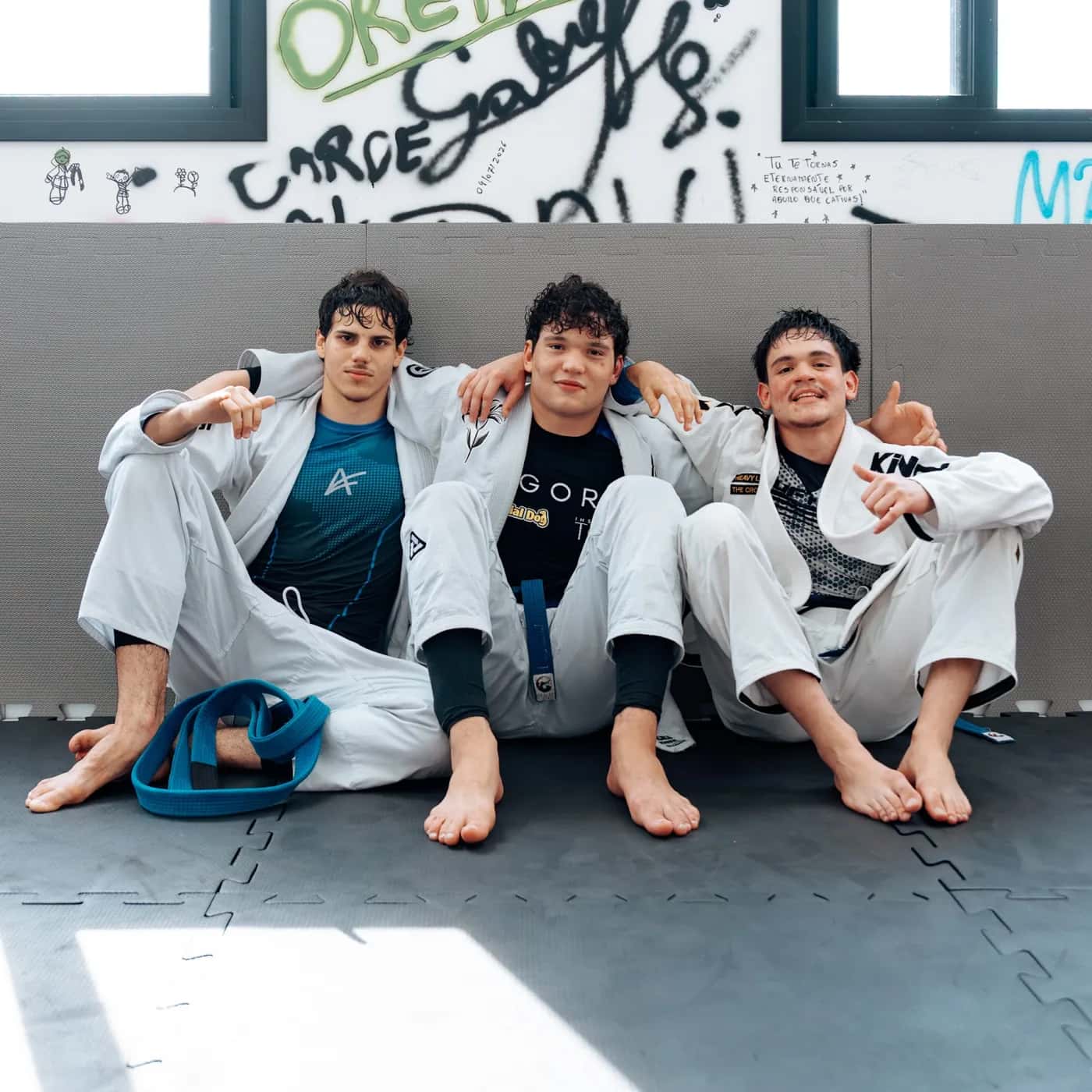 Três alunos de kimono sentados lado a lado no tatame, sorrindo